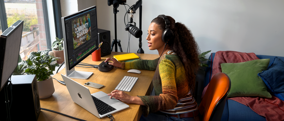 Mulher em casa, a trabalhar sentada à secretária com um MacBook Pro ligado a dois monitores externos, a usar auscultadores e a falar para um microfone