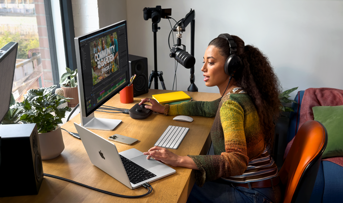 Mulher em casa, a trabalhar sentada à secretária com um MacBook Pro ligado a dois monitores externos, a usar auscultadores e a falar para um microfone