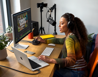 Mulher em casa, a trabalhar sentada à secretária com um MacBook Pro ligado a dois monitores externos, a usar auscultadores e a falar para um microfone